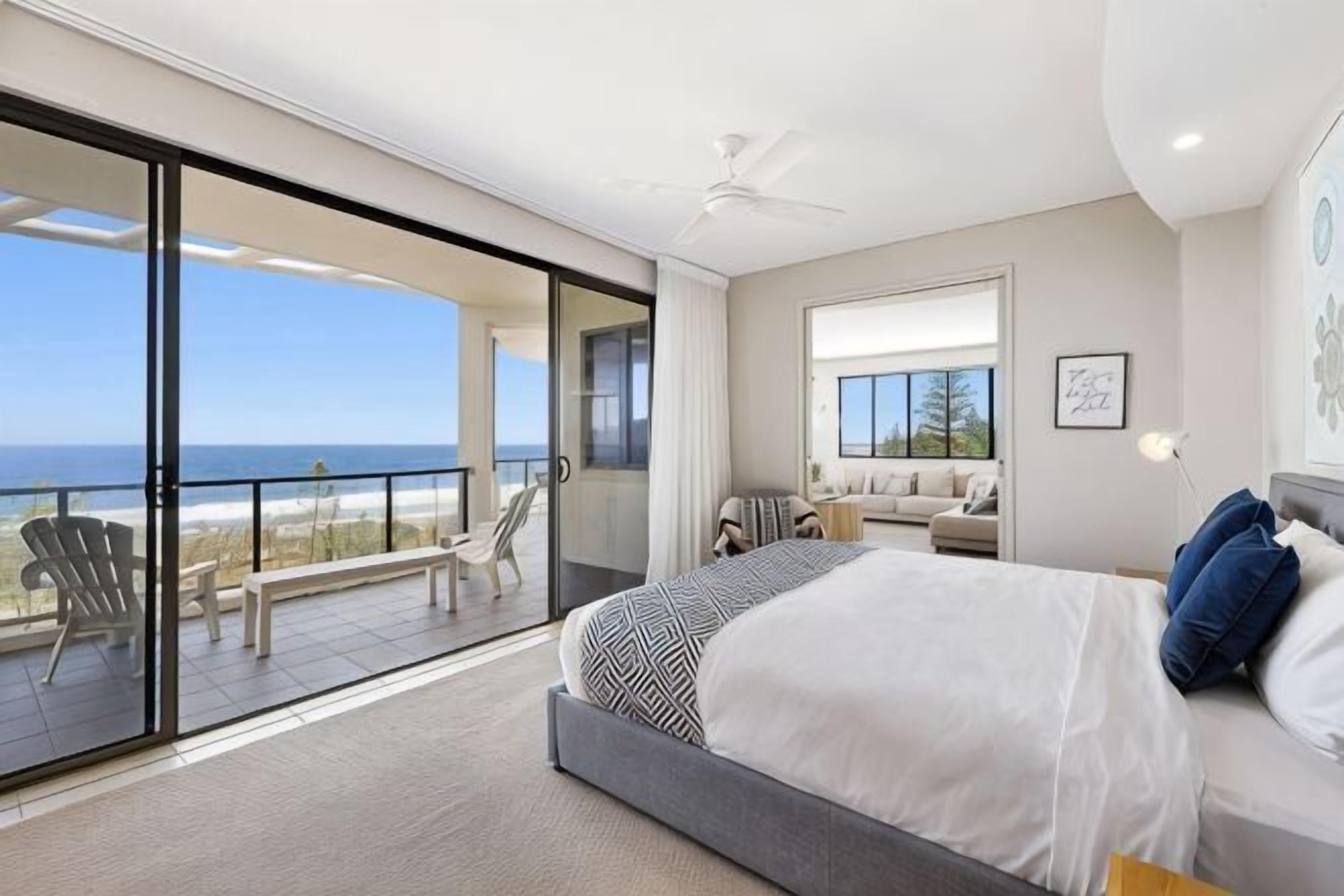 A bedroom with a large bed, a balcony with chairs, and a view of the ocean.