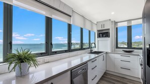 A modern kitchen with large windows, a dishwasher, and a view of the ocean. - Meridian Alex Beach Apartments (Alexandra Headland)