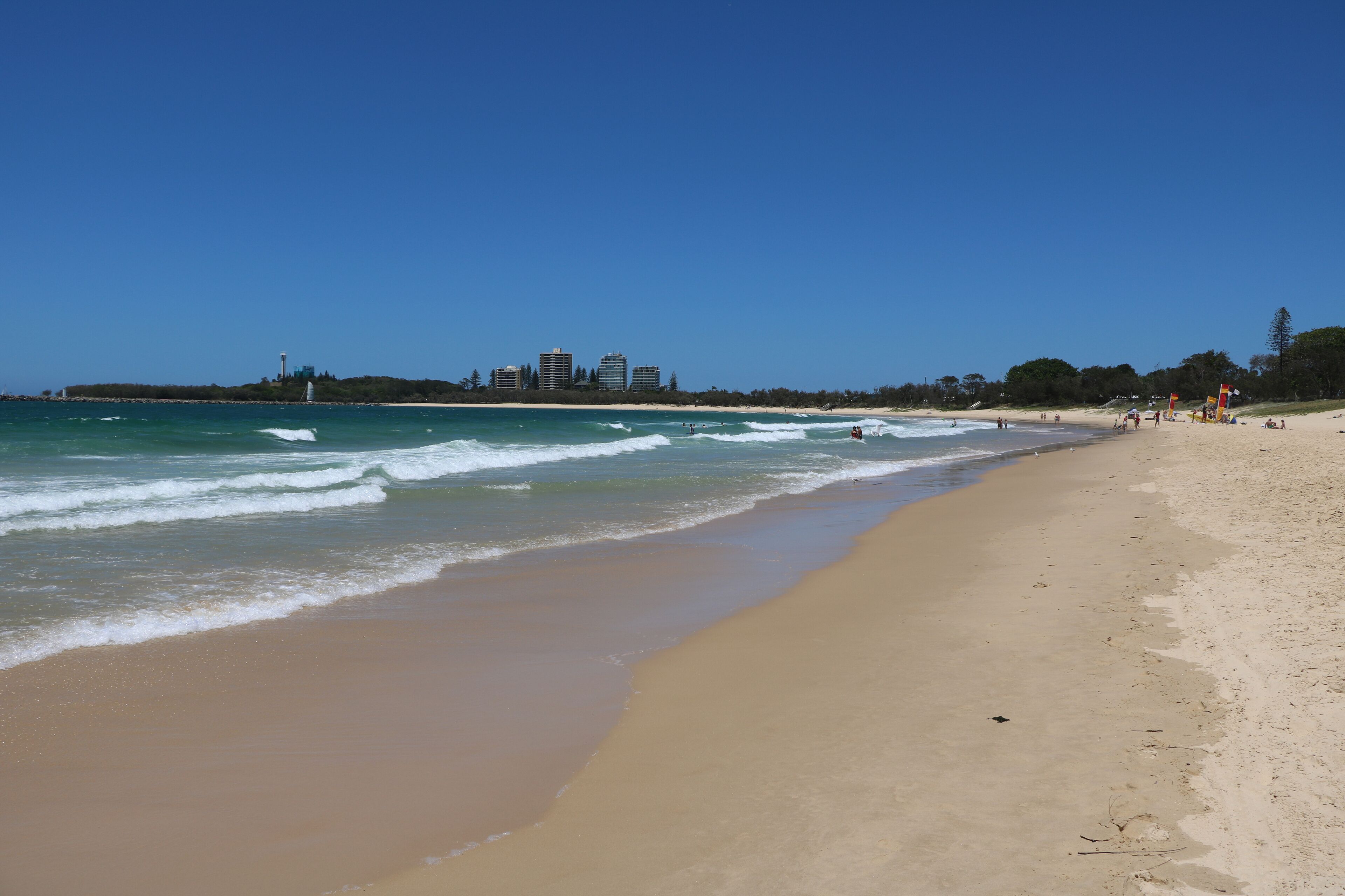 A wide sandy beach with gentle waves, a clear blue sky, and a distant city skyline.