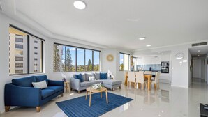 A modern living room with a blue sofa, a grey sofa, and a wooden coffee table. There is a kitchen area with white cabinets and a dining table. - Meridian Alex Beach Apartments (Alexandra Headland)