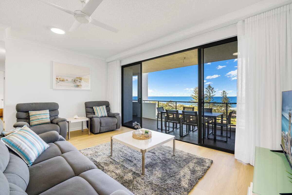 Una sala de estar moderna con un sofá gris, dos sillones, una mesa de centro y un amplio ventanal corredero que da a un patio exterior con vista al océano.
