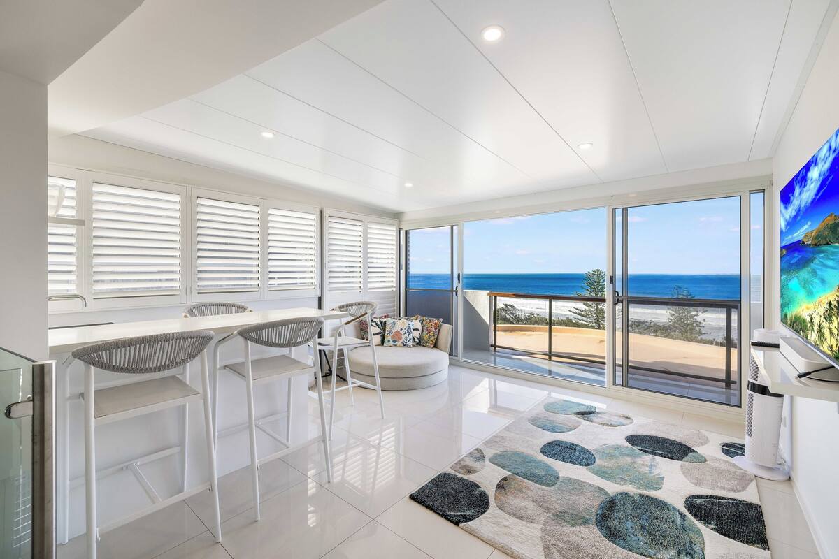 Una cocina moderna con armarios blancos, un comedor con mesa y sillas redondas, y ventanas amplias con vista al océano.