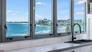 A kitchen with a large window overlooking a beach and cityscape. - Meridian Alex Beach Apartments (Alexandra Headland)