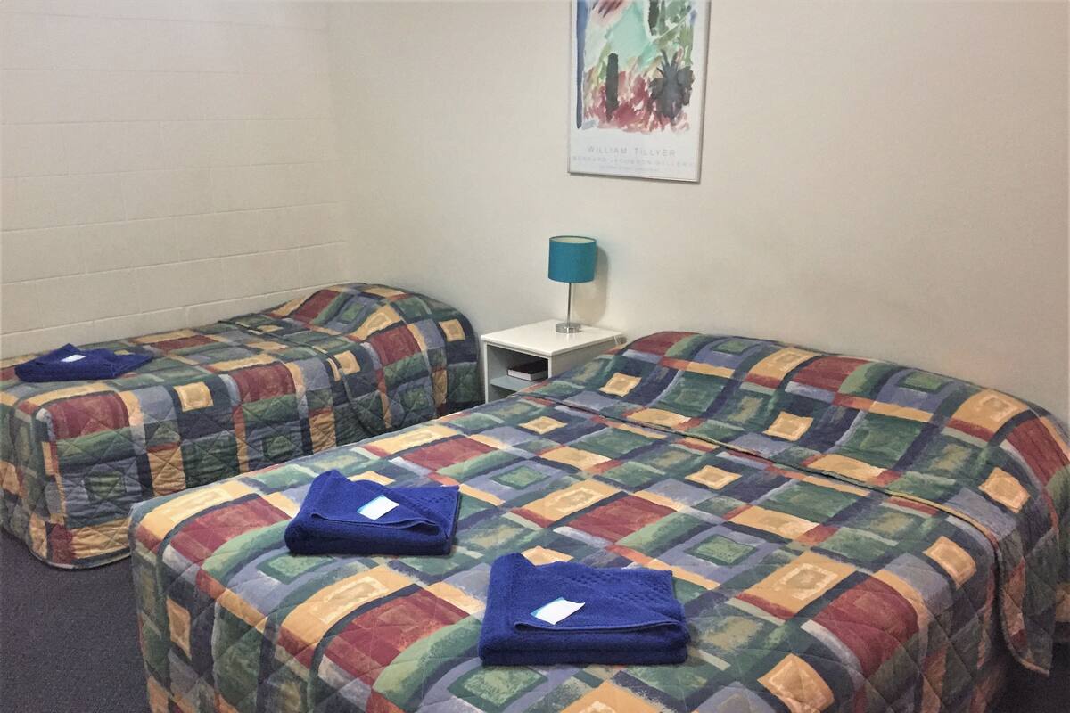 Una habitación con dos camas individuales y una cama más grande, cada una con colchas de colores. Hay una mesita con una lámpara y un cuadro enmarcado en la pared.