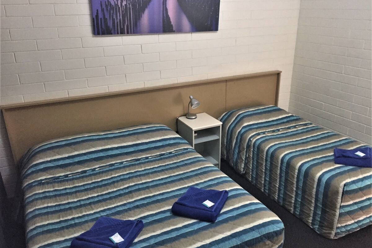 Habitación de hotel con dos camas, cada una con ropa de cama a rayas y una almohada. Hay una mesita de noche con una lámpara y un cuadro en la pared sobre las camas.