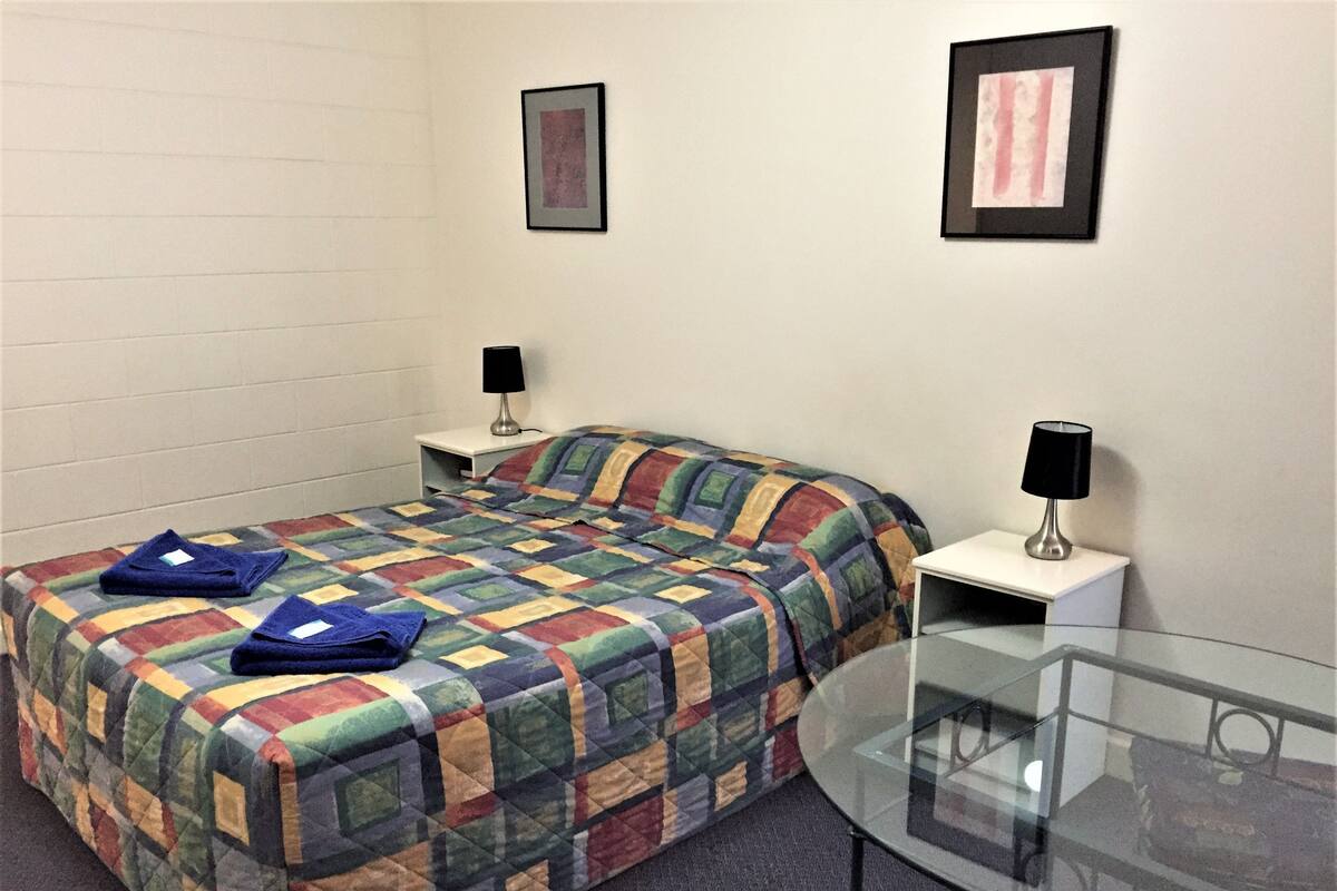 Una cama individual con un edredón colorido, dos mesitas de noche con lámparas, y una mesa de vidrio en la esquina.