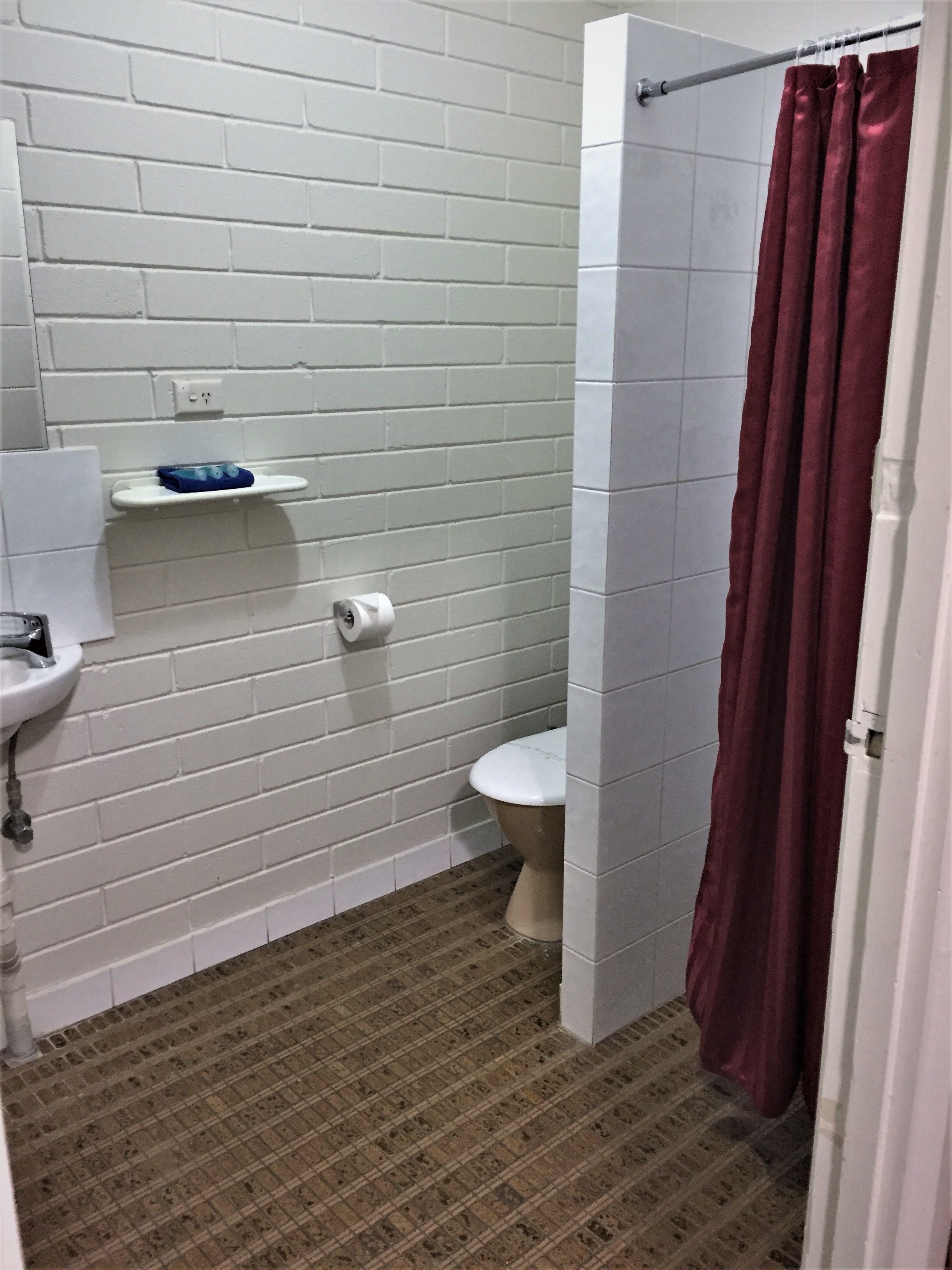 Un baño con pared de ladrillo blanco, un inodoro blanco, un lavabo con una toalla azul y una cortina de ducha.