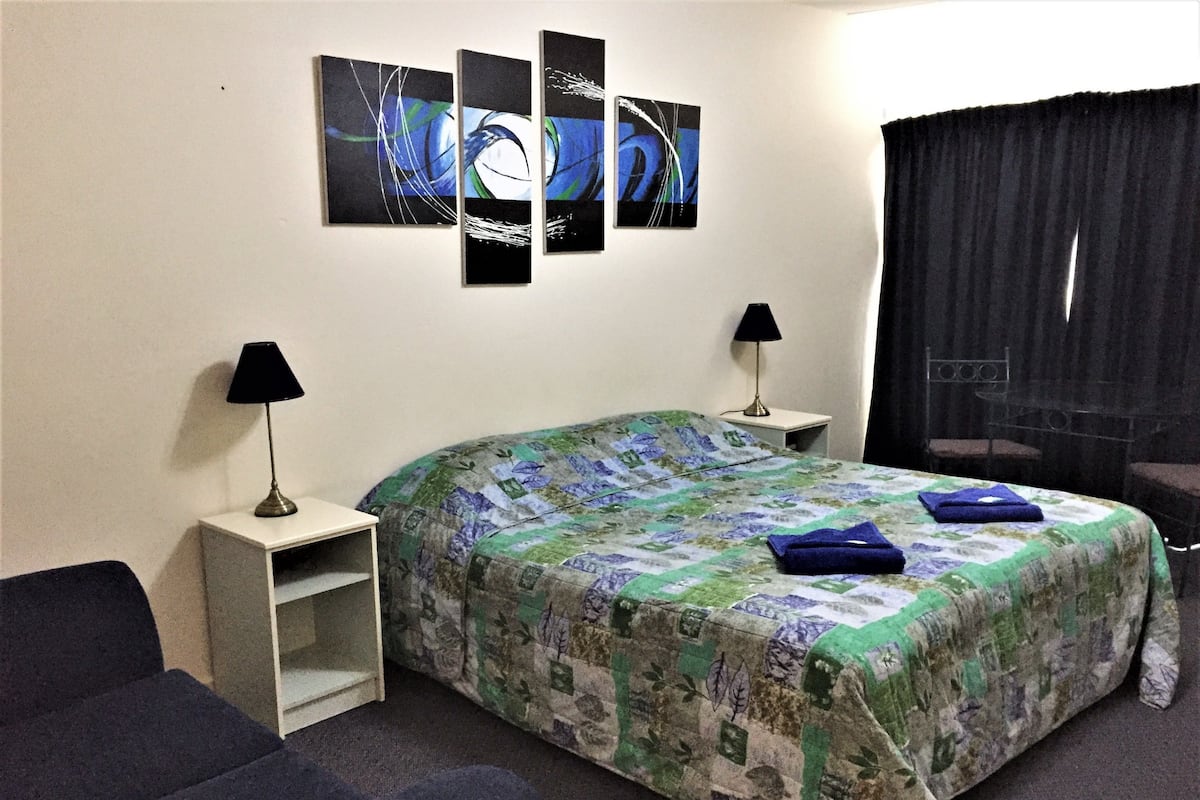 Una habitación de hotel con cama, dos mesitas de noche, lámpara, silla y ventana con cortinas.