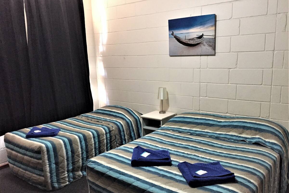 Habitación de hotel con dos camas, una mesita de noche, una lámpara y un cuadro de un barco en la pared.