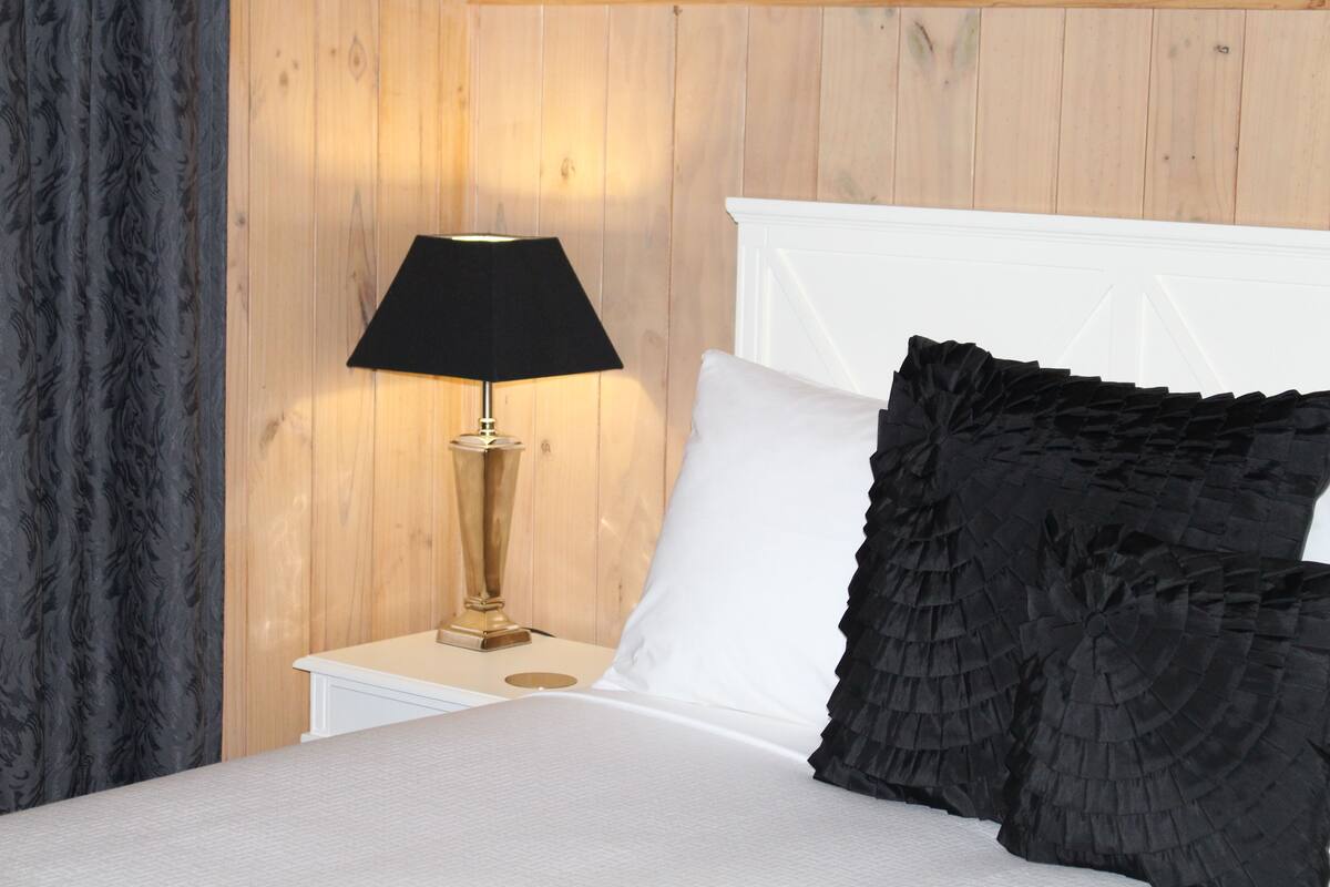 A neatly made bed with a white headboard, a black ruffled pillow, and a gold lamp on a bedside table against a wooden wall.