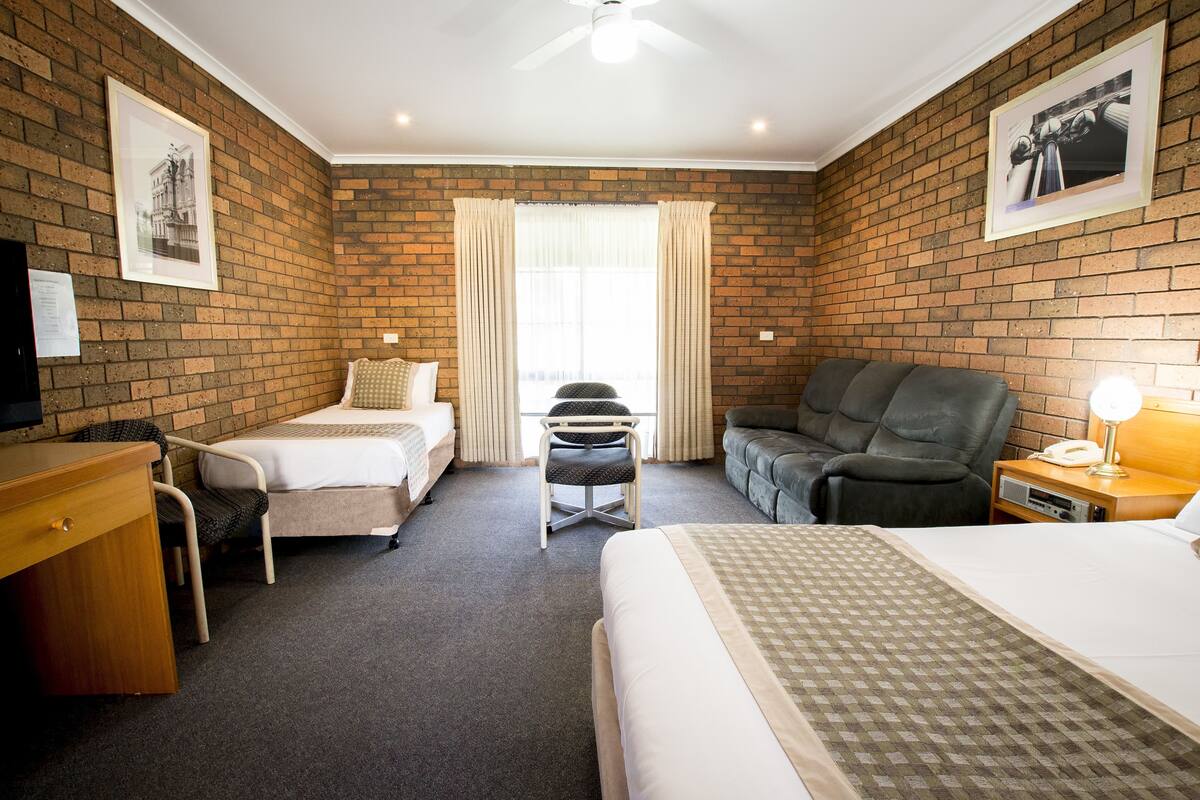 A hotel room with a brick wall, two beds, a desk, a chair, a sofa, and a window with curtains.
