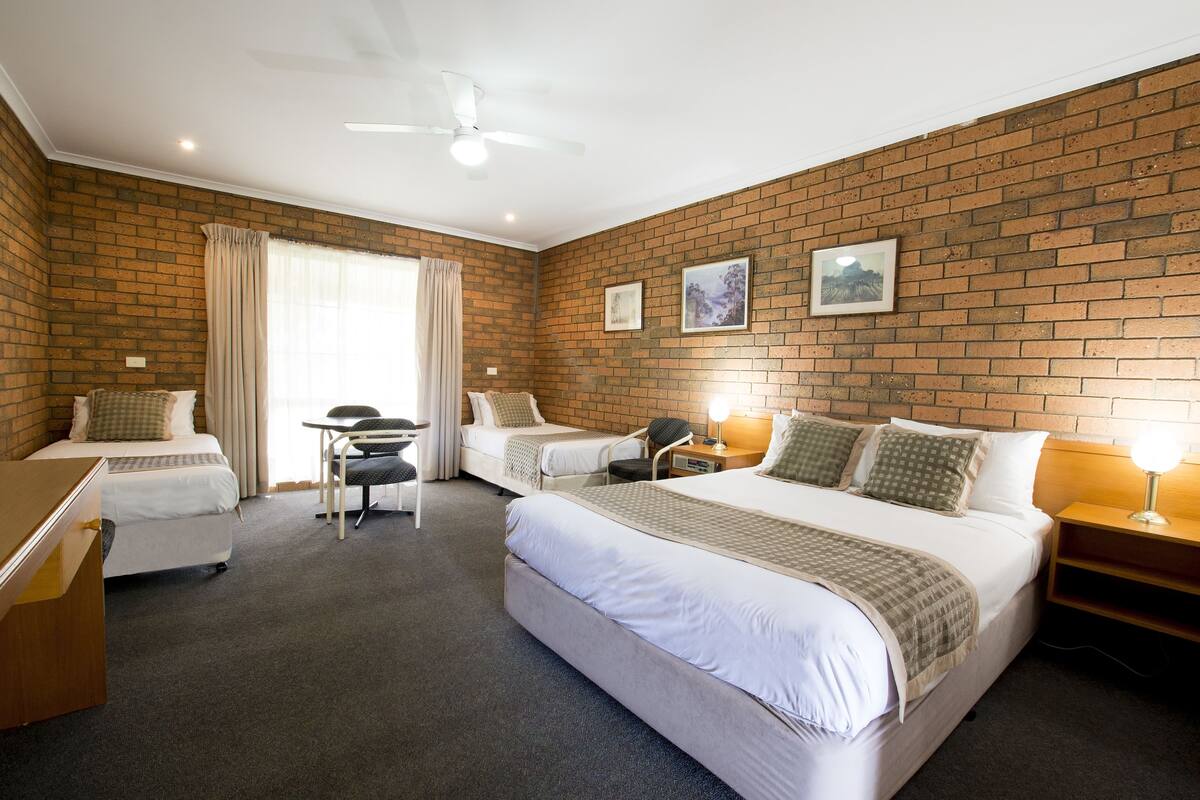 A hotel room with two single beds, a desk, a chair, a small table, and a nightstand with a lamp.