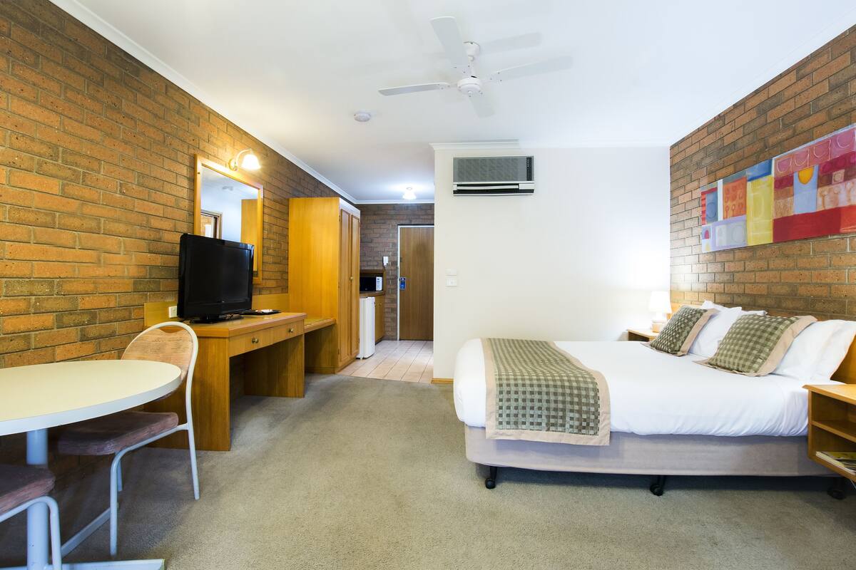 A hotel room with a brick wall, a bed with a checkered blanket, a desk with a television, and a ceiling fan.