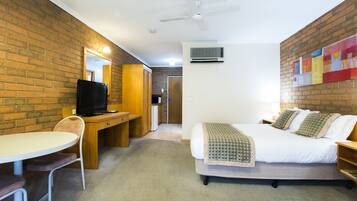 A hotel room with a brick wall, a bed with a checkered blanket, a desk with a television, and a ceiling fan.