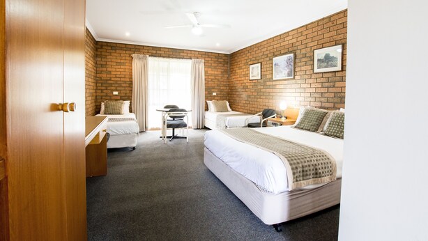 A hotel room with two single beds, a desk, a chair, a ceiling fan, and a brick wall with framed pictures. - Sportslander Motor Inn (Moama)