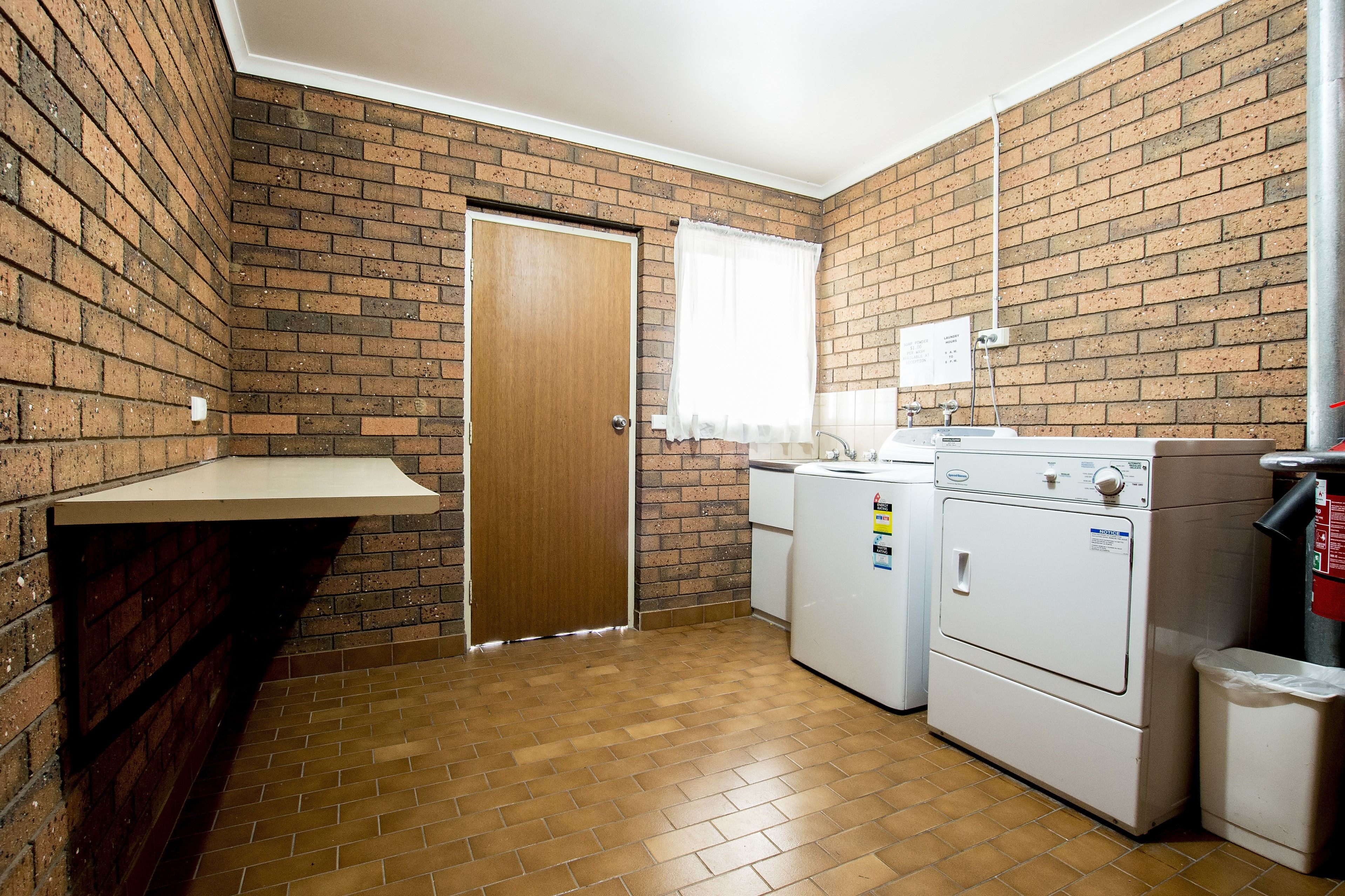 A laundry room with a brick wall, a wooden door, a window with white curtains, a washing machine, a dryer, and a trash can.