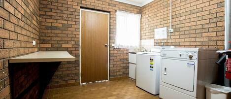 Una sala de lavandería con pared de ladrillo, una puerta de madera, una ventana con cortinas blancas, una lavadora, una secadora y un bote de basura.