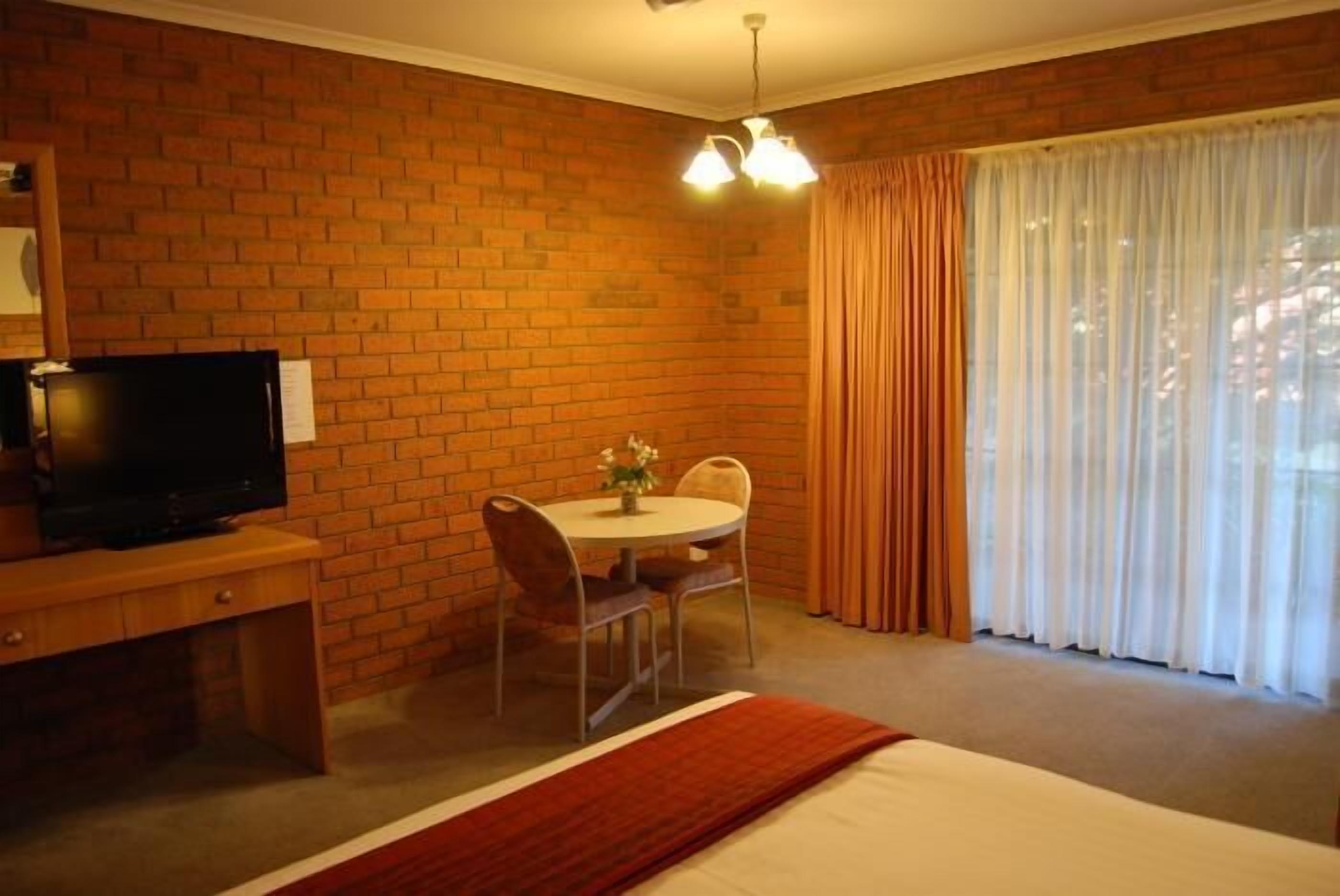 Quarto de hotel com parede de tijolos, televisão em escrivaninha de madeira, mesa redonda com cadeiras, cama com edredom branco e janela com cortinas.