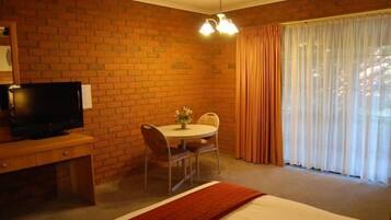 Quarto de hotel com parede de tijolos, televisão em escrivaninha de madeira, mesa redonda com cadeiras, cama com edredom branco e janela com cortinas.
