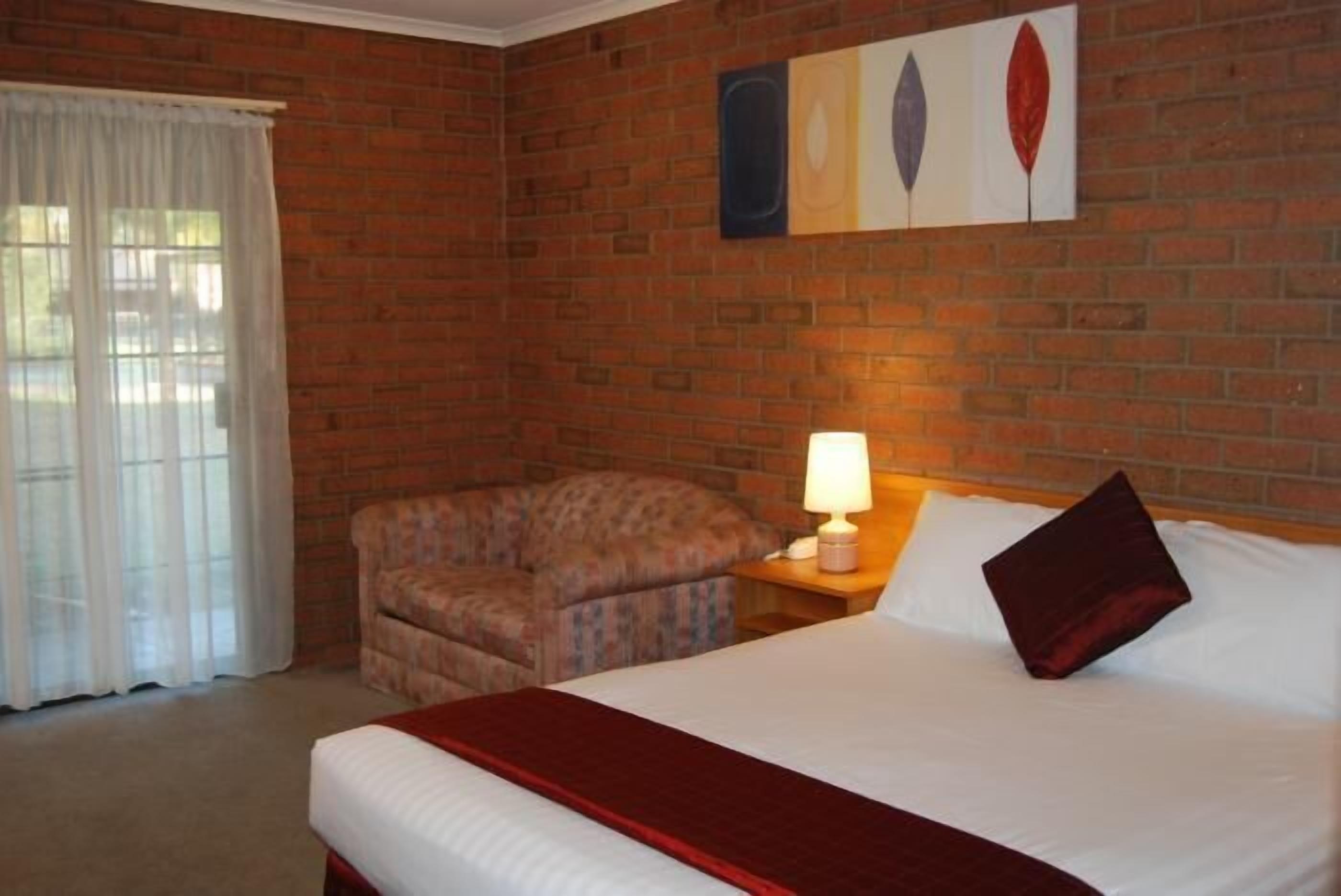 A hotel room with a brick wall, a bed with a red bedspread, a chair, a nightstand with a lamp, and a window with white curtains.