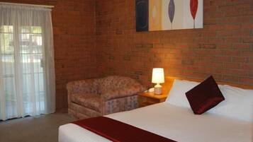 A hotel room with a brick wall, a bed with a red bedspread, a chair, a nightstand with a lamp, and a window with white curtains.