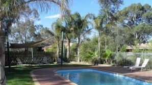 A rectangular swimming pool with a brick border, surrounded by trees and a shaded area with lounge chairs. - Sportslander Motor Inn (Moama)