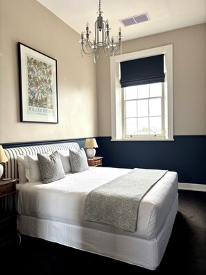 A bedroom with a large bed, a chandelier, a framed picture on the wall, a window with a dark valance, and a nightstand with a lamp.