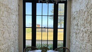 A room with a round wooden table and wicker chairs, a chandelier, and a large arched window with a view of a field and buildings. - The Customs House Port MacDonnell (Port Macdonnell)