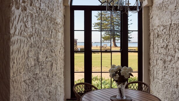 A wooden table with a vase of white flowers, set against a textured wall with a large window overlooking a grassy area and a tree. - The Customs House Port MacDonnell (Port Macdonnell)