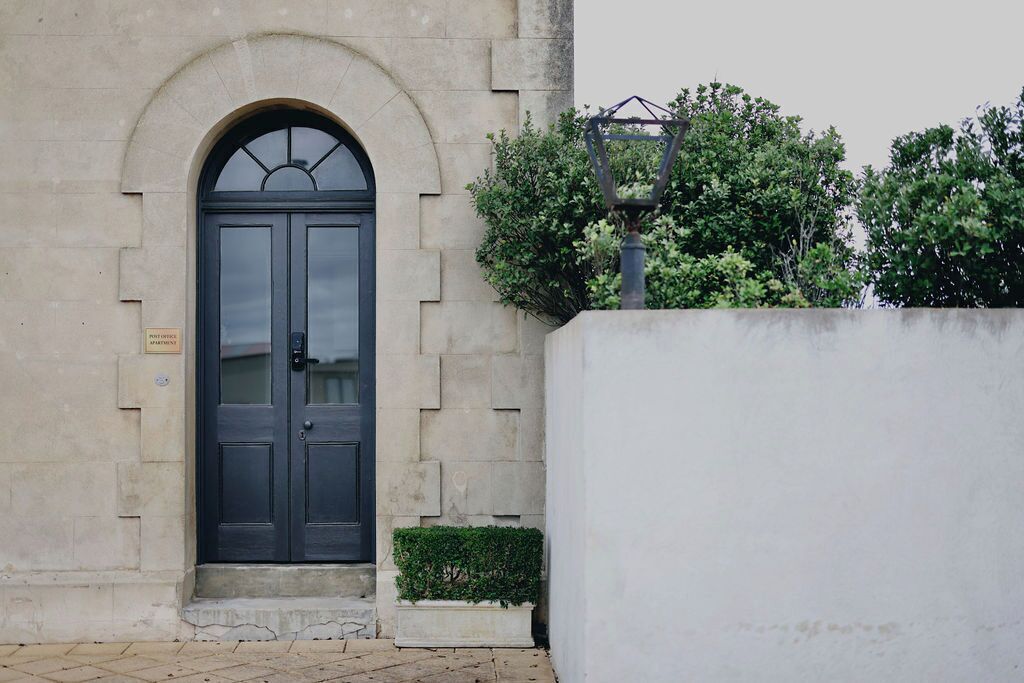 A building with a dark arched entrance door, a small step leading up to it, and a white wall with a planter of greenery beside it.