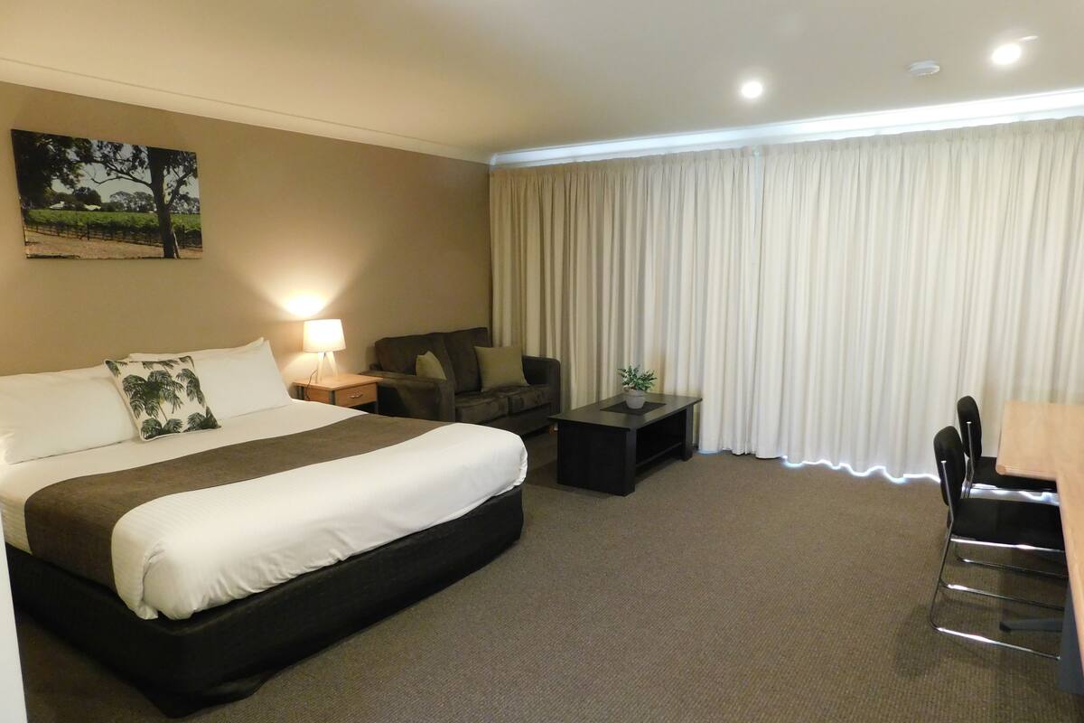 Habitación de hotel con cama, escritorio, silla, una mesita y ventana con cortinas.