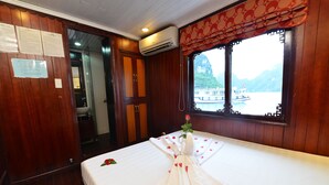 A room with wooden paneling, a bed with a white bedspread and rose petals, a window with a view of a boat and mountains, and an air conditioning unit. - Imperial Classic Cruise Halong (Tuan Chau)
