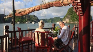 A deck with wooden chairs and tables, overlooking a body of water with boats and greenery in the background. - Imperial Classic Cruise Halong (Tuan Chau)