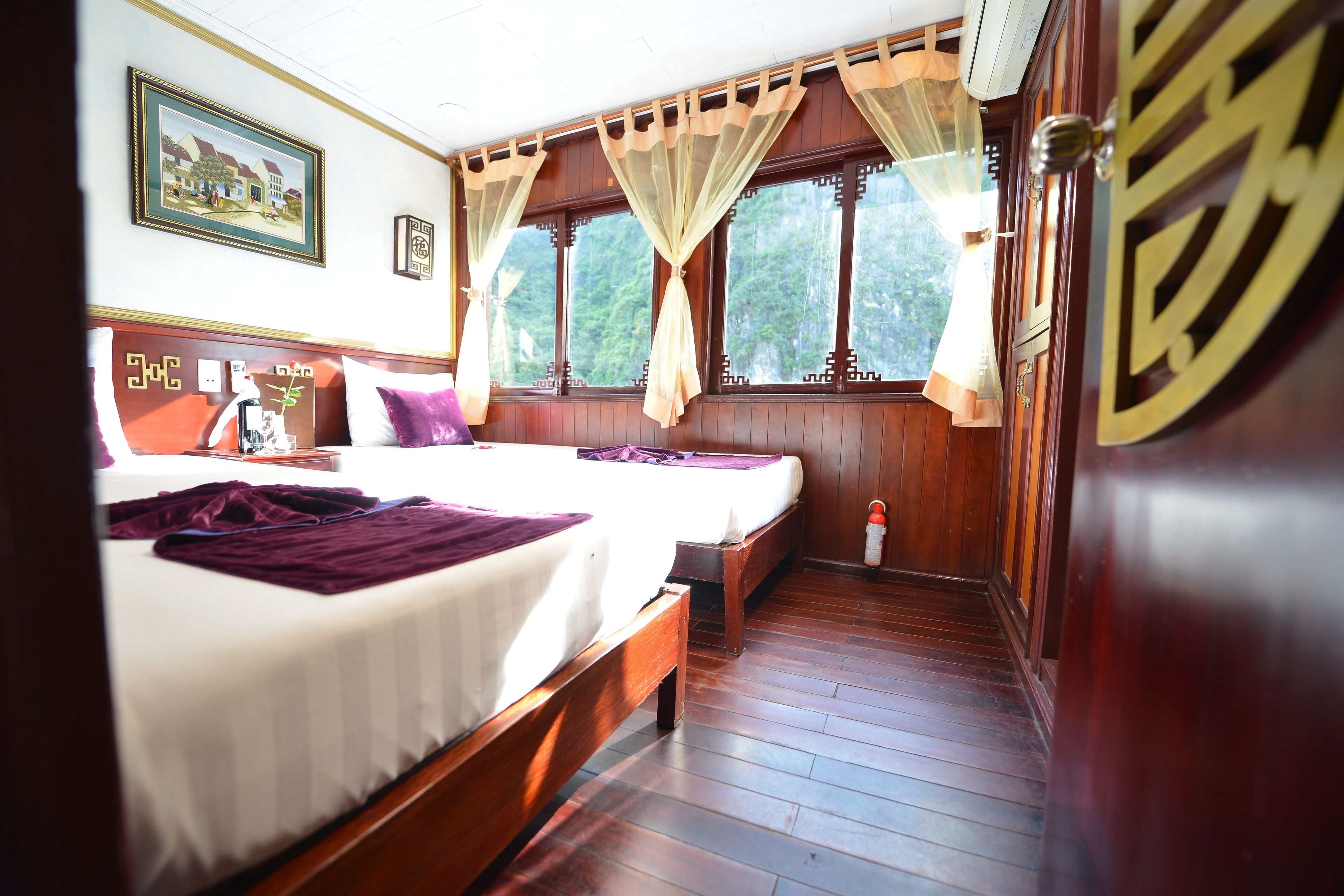 A hotel room with two beds, wooden flooring, a large window with curtains, and a framed picture on the wall.