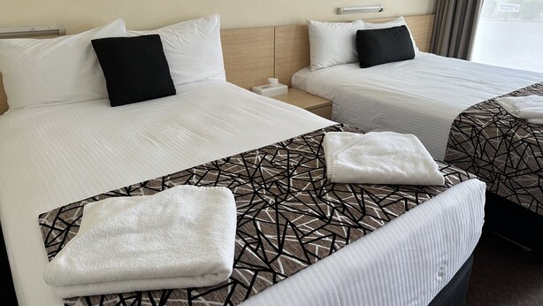 A hotel room with two beds, each with white bedding and black pillows. There are towels on the beds and a nightstand between them. - City Centre Motel (Kempsey)