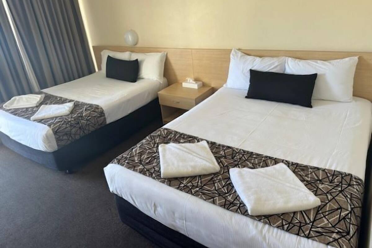 Two twin beds with patterned bedding and white pillows, a bedside table with a lamp, and a window with curtains.