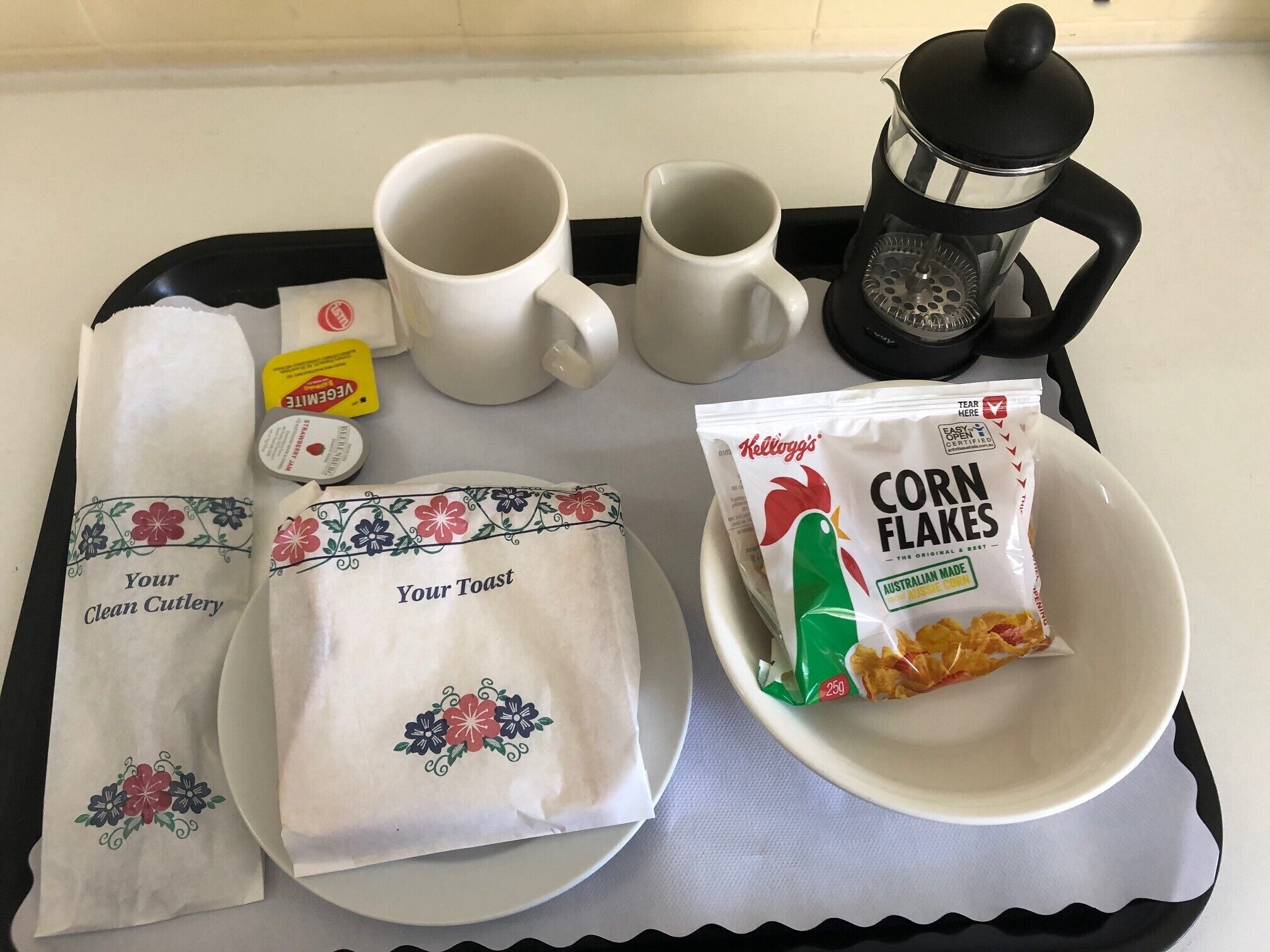 A tray with a coffee maker, two mugs, a packet of corn flakes, a packet of sugar, a packet of tea, and two cloth napkins with floral designs.