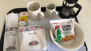 A tray with a coffee maker, two mugs, a packet of corn flakes, a packet of sugar, a packet of tea, and two cloth napkins with floral designs.