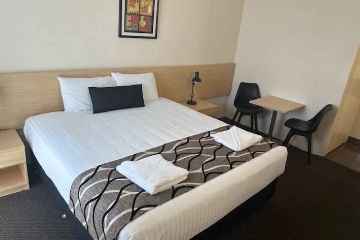 A hotel room with a bed, a desk, a chair, a nightstand, and a framed picture on the wall.
