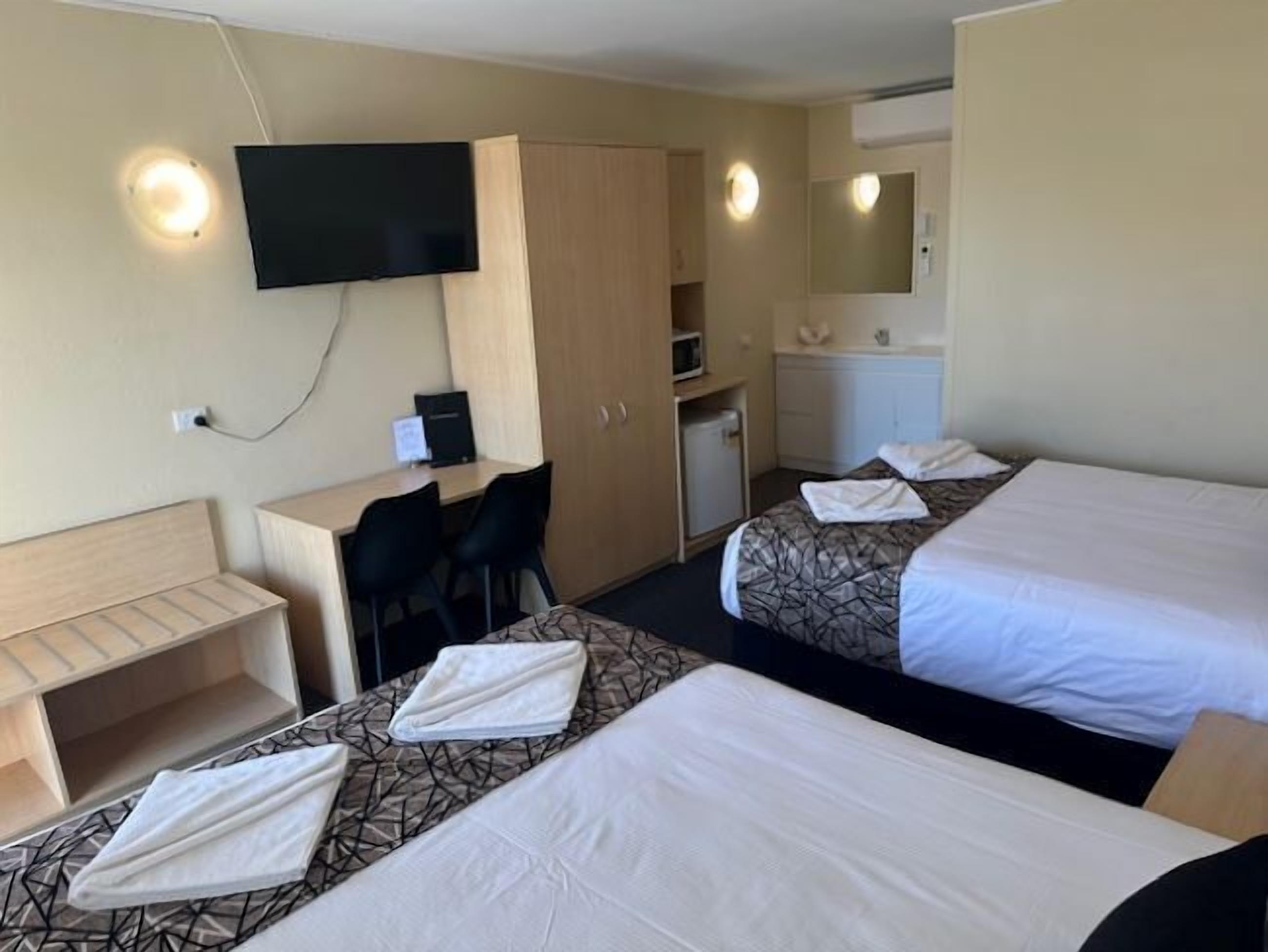 A hotel room with two beds, a desk, a chair, a television, and a small kitchenette.