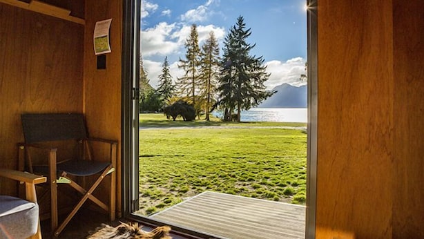 A room with a large window offering a view of a grassy area, trees, and a body of water. - The Camp - Lake Hawea (Lake Hawea)