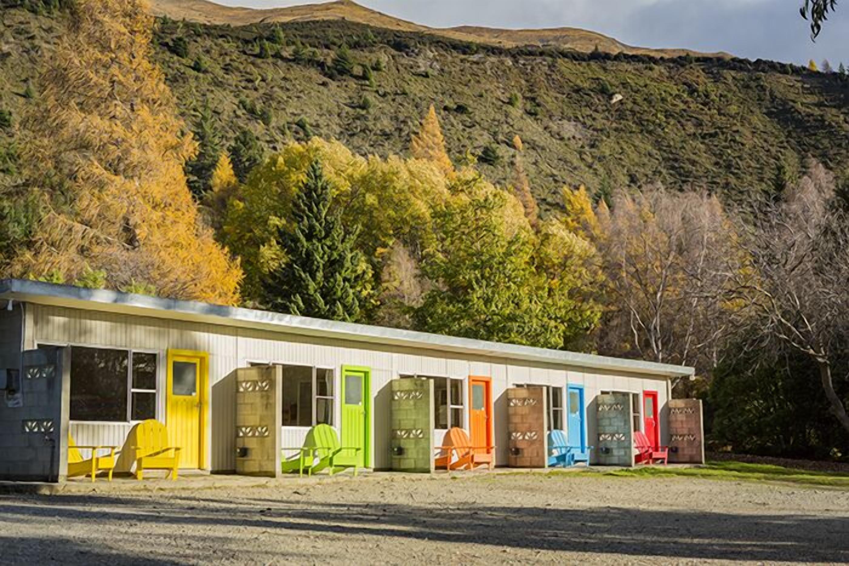 A row of colorful cabins with individual doors, set against a backdrop of a hilly landscape.