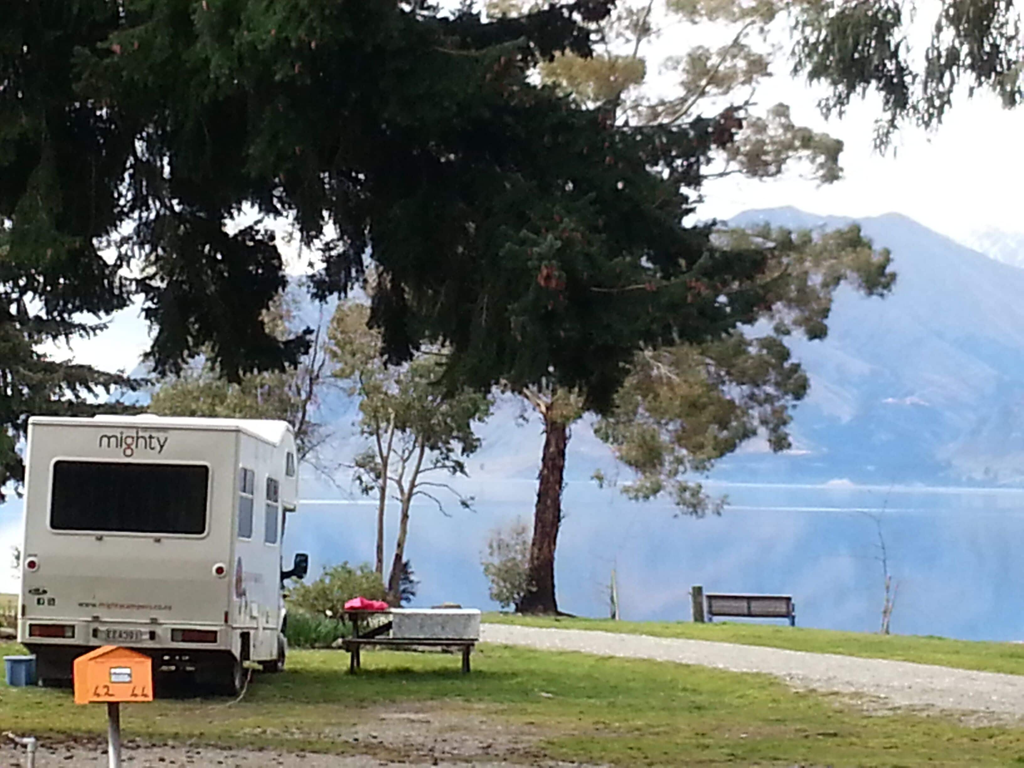 Una caravana llamada 'mighty' estacionada cerca de un lago, con un banco y una mesa al frente.
