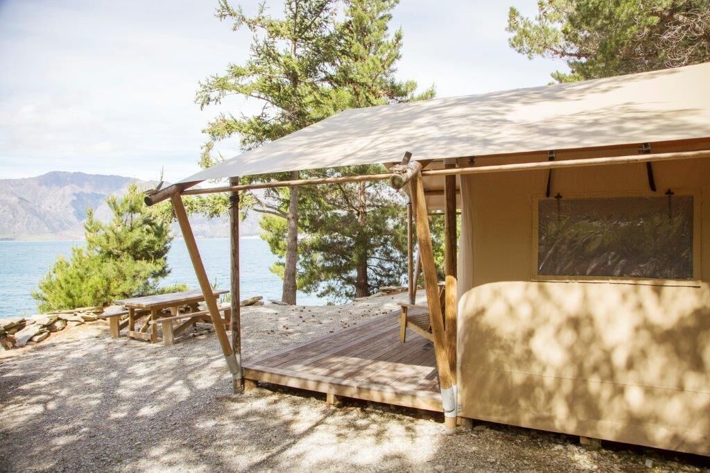 A tented camping area with a wooden deck, picnic tables, and a view of a lake and mountains.