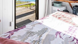 A bedroom with a bed, a desk, a chair, a window with a view of a lake and mountains, and a fire extinguisher on the wall. - The Camp - Lake Hawea (Lake Hawea)