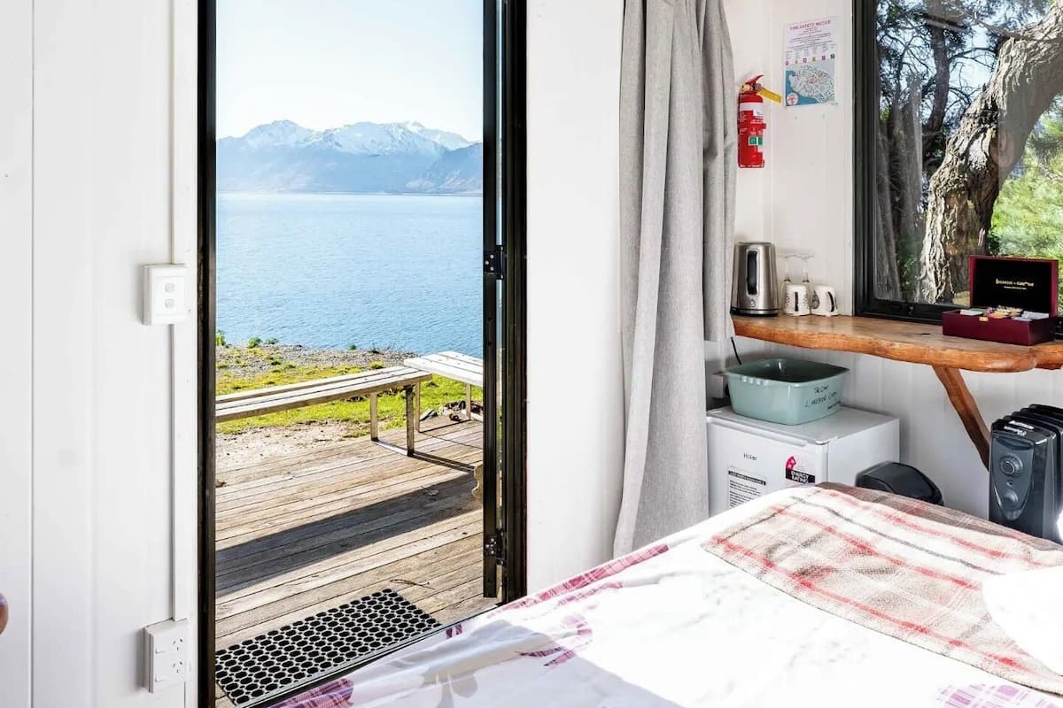 Un dormitorio con cama, escritorio, silla, ventana que ofrece vistas a un lago y montañas, y un extintor de incendios en la pared.
