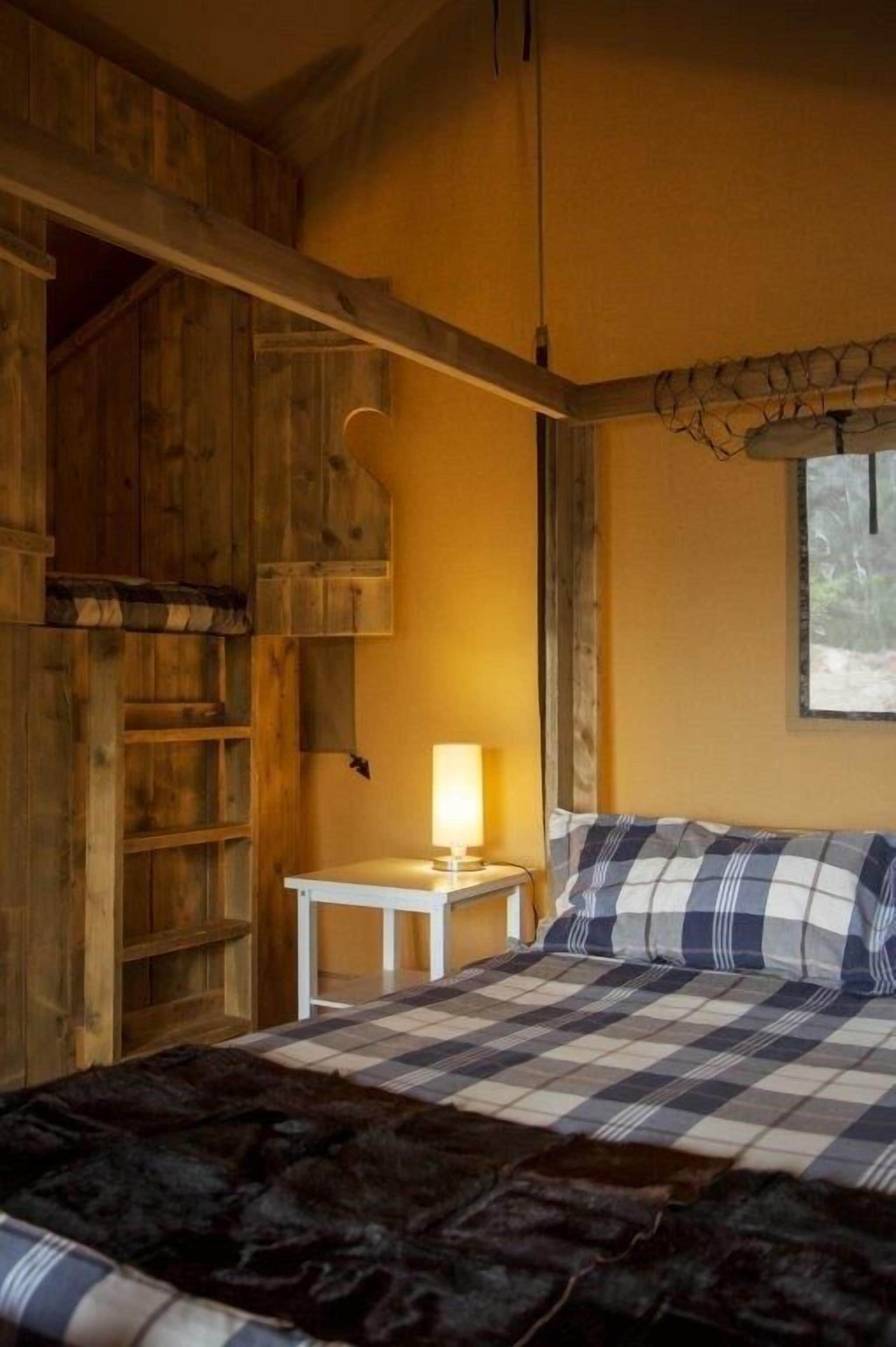 A cozy room with a bed, a small table with a lamp, wooden shelves, and a window.