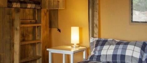 A cozy room with a bed, a small table with a lamp, wooden shelves, and a window.
