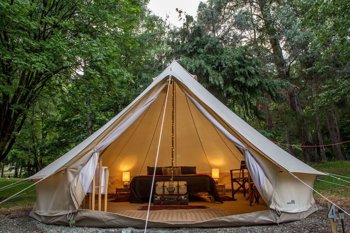 Una carpa grande de color beige con una cama y dos lámparas en su interior, rodeada de árboles.