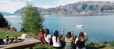 A group of people sitting on a wooden bench overlooking a lake with mountains in the background.